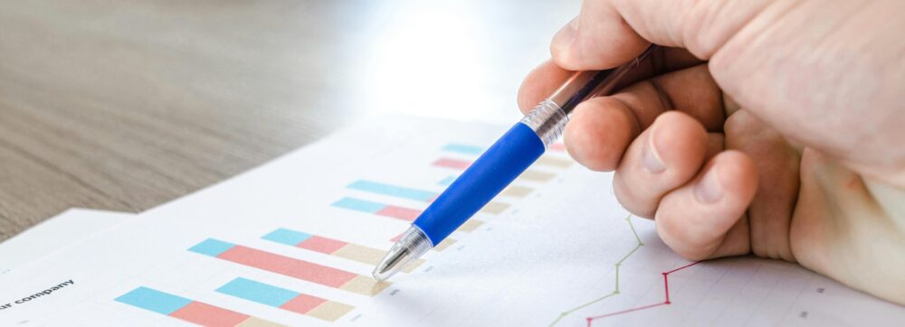 A close-up of a hand with a pen analyzing data on colorful bar and line charts on paper.